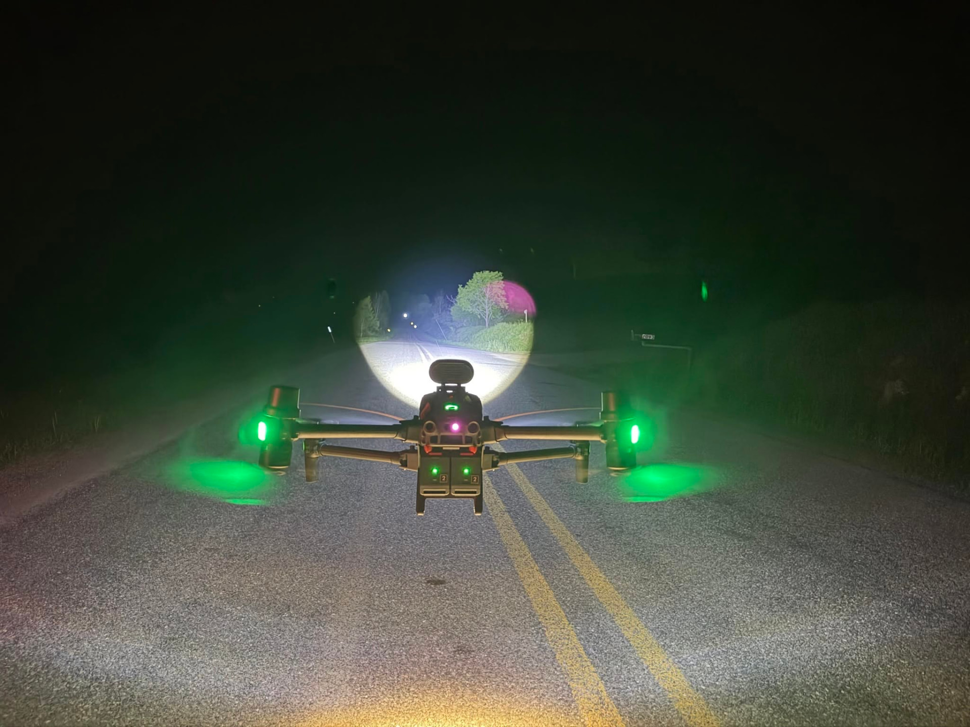 Thermal search drone hovering over a rural road at night with green navigation lights and a white spotlight beam